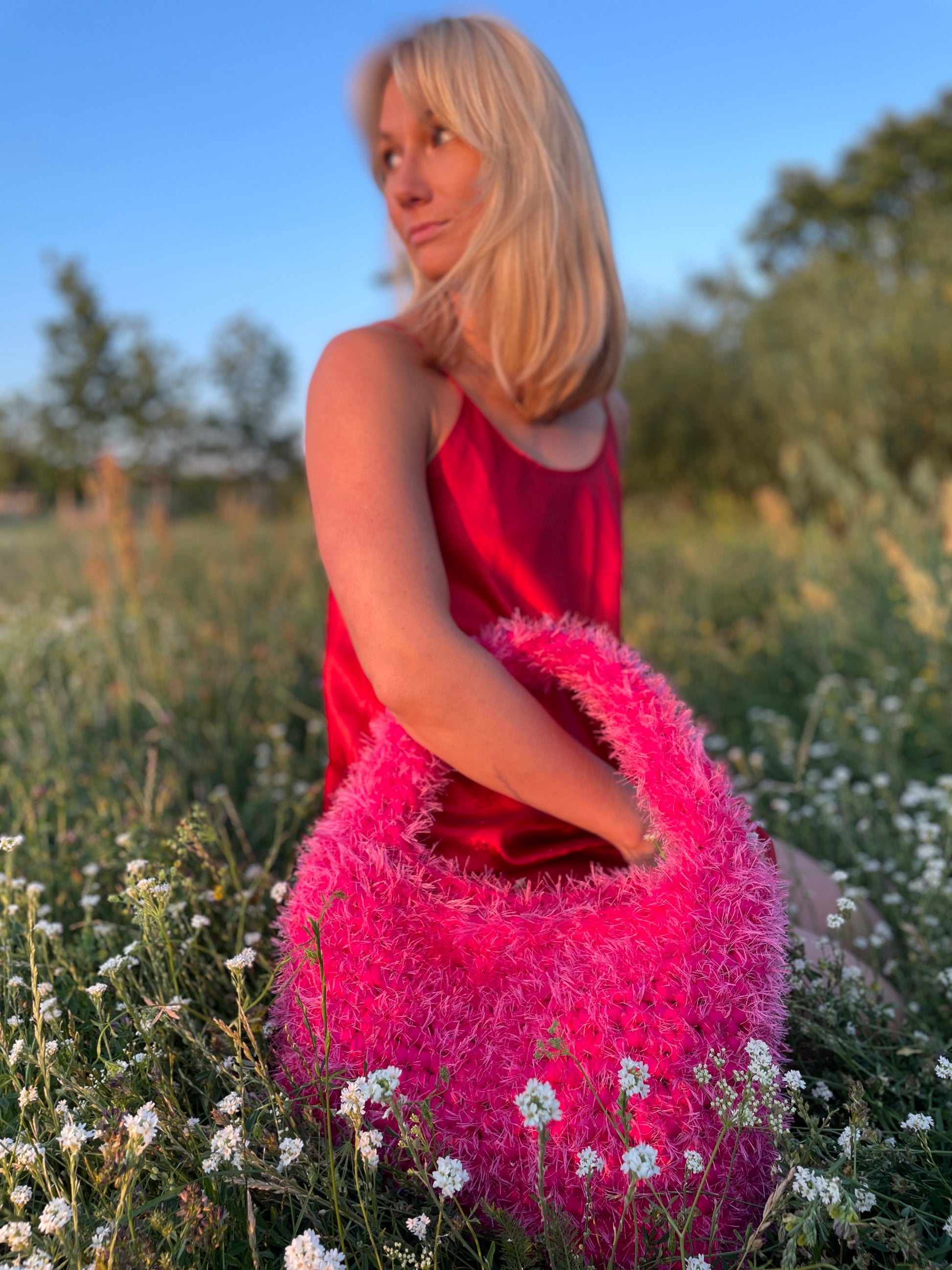 A blond model wearing a fluffy hot pink crochet bag with a satin red dress in the park with white flowers in the foreground. It is a large fluffy pink bag, pink tote bag.