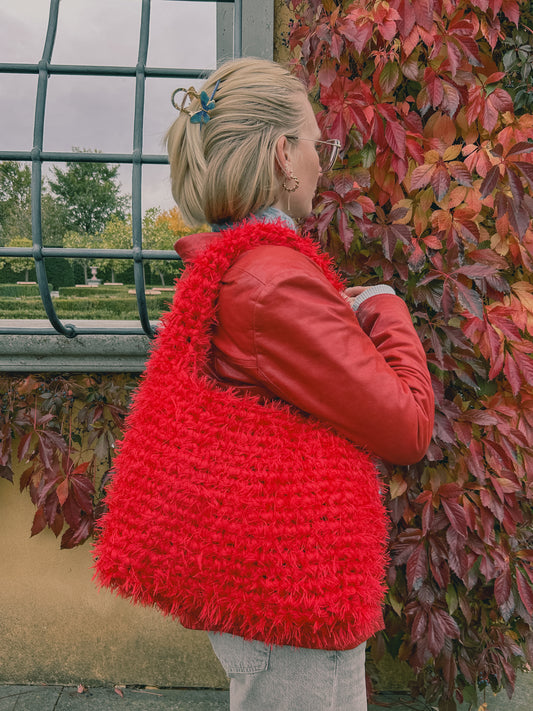A person wearing a red crochet bag with fluffy detailing, standing by a window with a view of autumn leaves.
