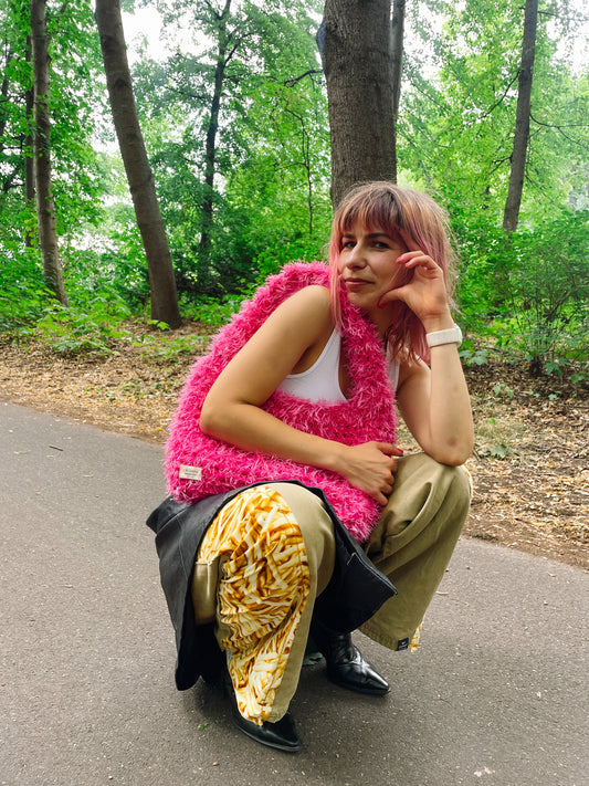 A person sitting on a road wearing a fluffy hot pink crochet bag with a white tank top and patterned pants. It is a large fluffy pink bag, pink tote bag.