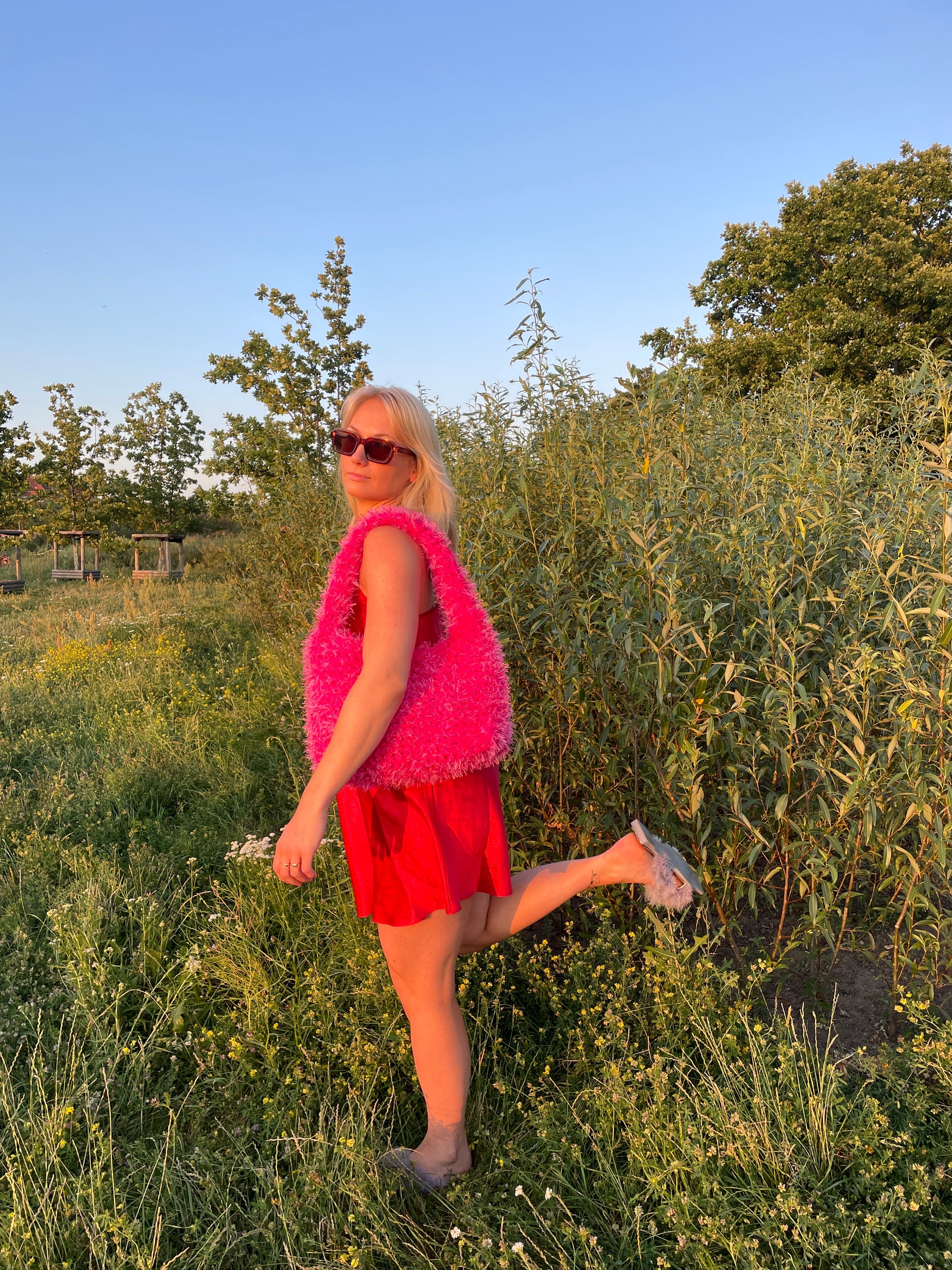 A blond model wearing a red satin mini dress carrying a fluffy hot pink crochet bag in the park with green grassy background. It is a large fluffy pink bag, pink tote bag.