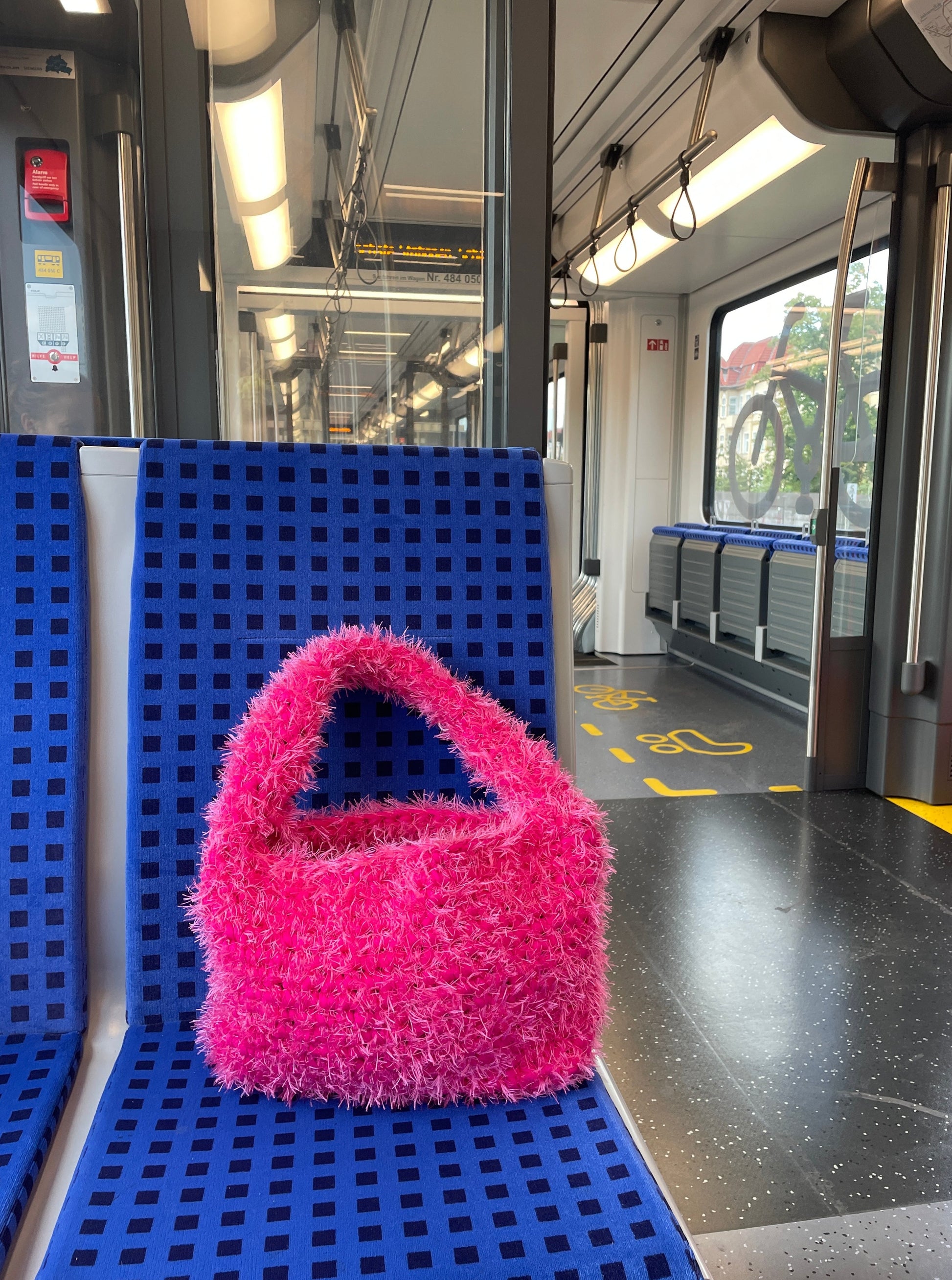 Product image of a fluffy hot pink crochet bag on a train seat. It is a large fluffy pink bag, pink tote bag, on a blue train seat.