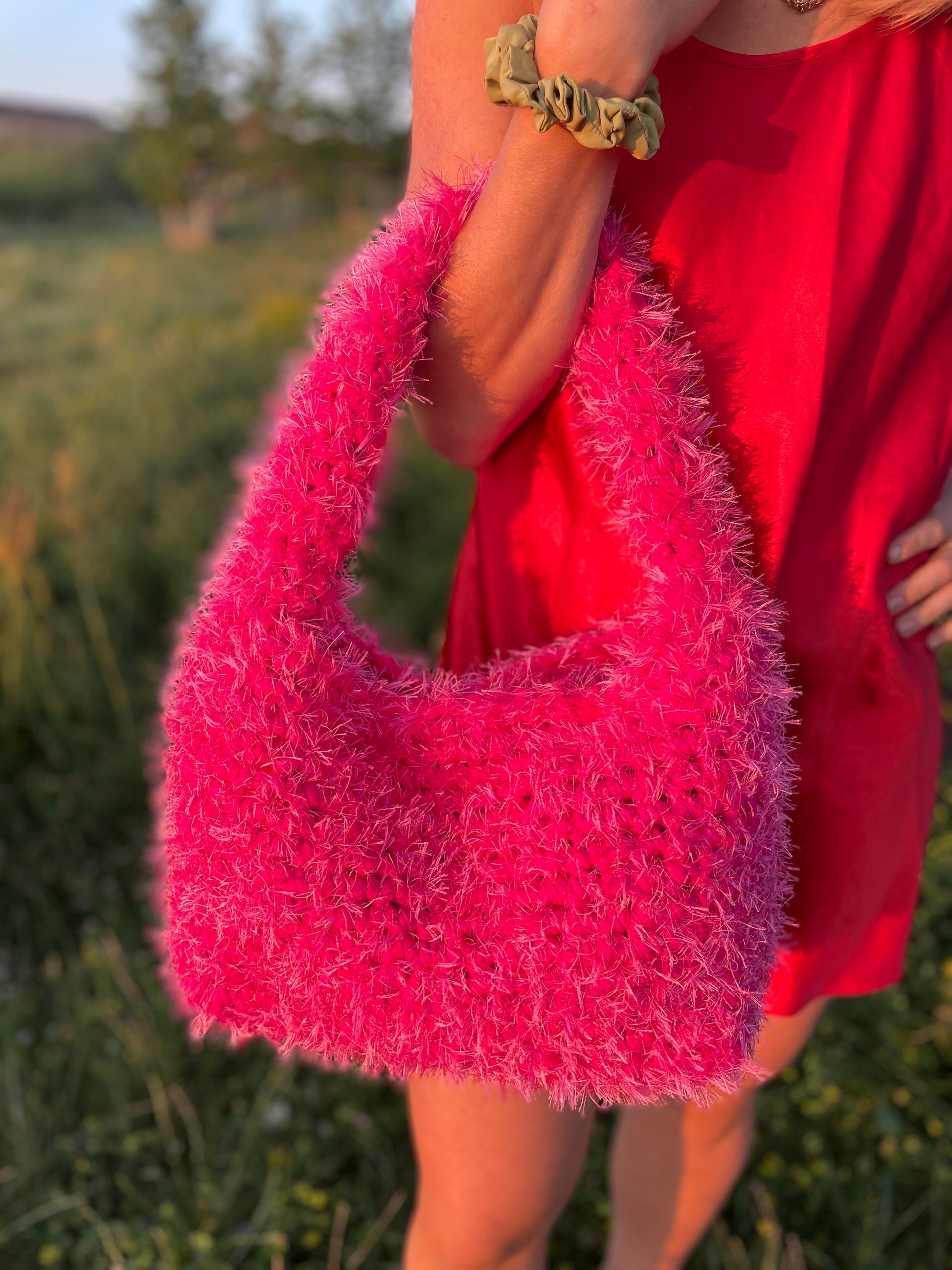 A person wearing a red satin mini dress carrying a fluffy hot pink crochet bag in the park with green grassy background. It is a large fluffy pink bag, pink tote bag.