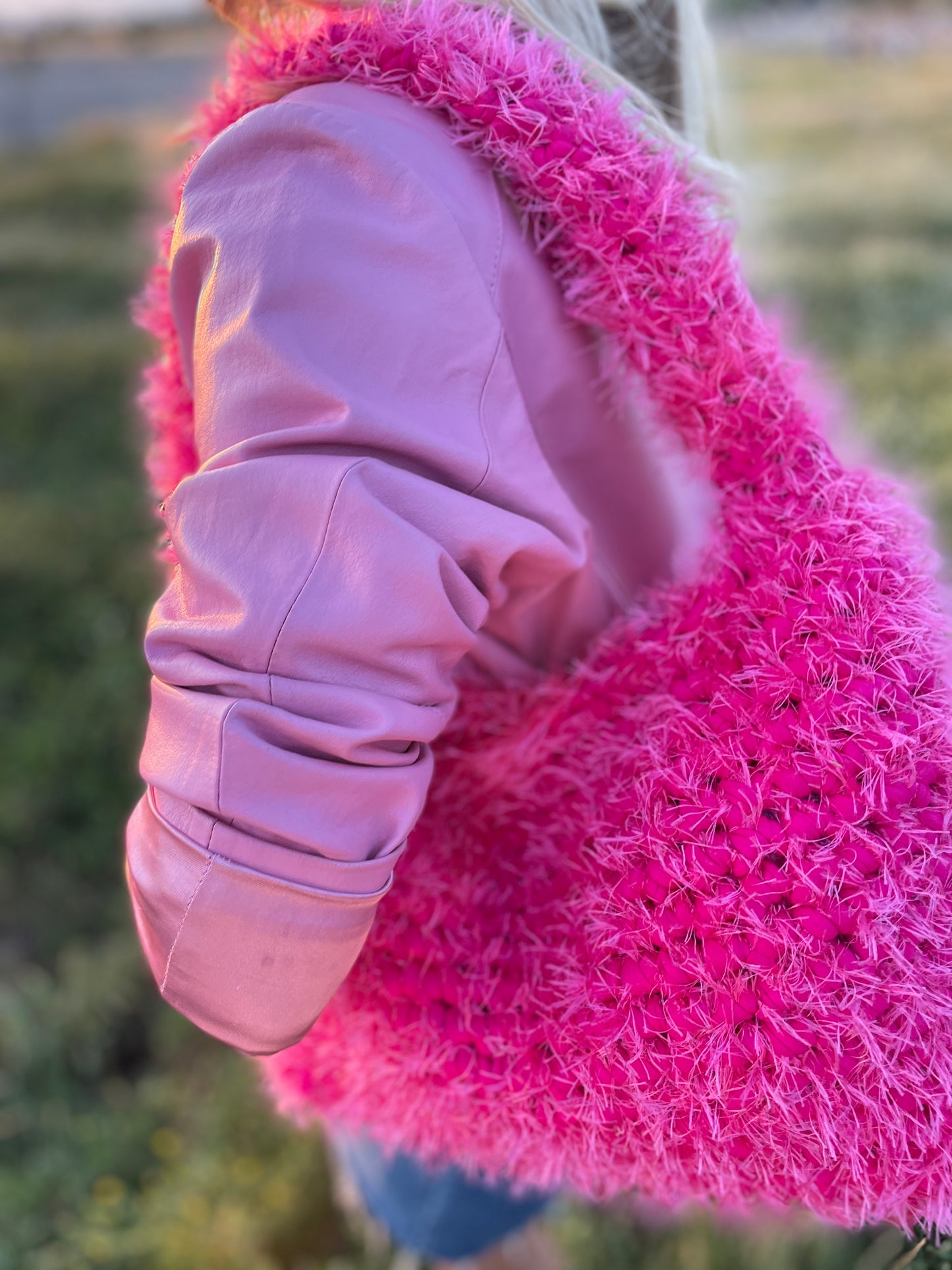 A product close up image of a fluffy hot pink crochet bag with a baby pink leather jacket, in the park. It is a large fluffy pink bag, pink tote bag.
