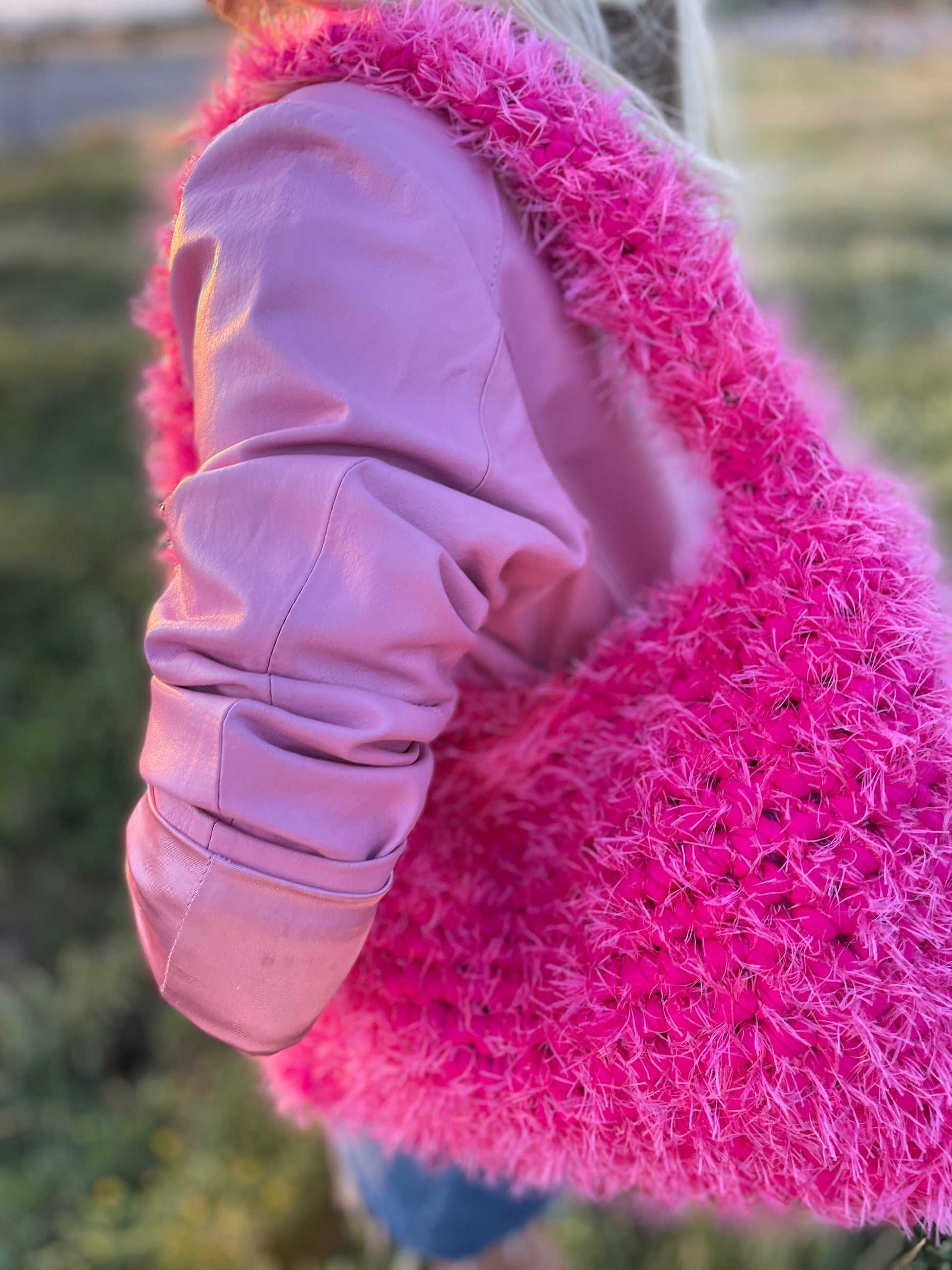A product close up image of a fluffy hot pink crochet bag with a baby pink leather jacket, in the park. It is a large fluffy pink bag, pink tote bag.