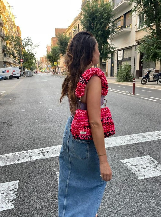 A person standing on a street wearing a pink and red crochet bag with black detailing, denim shorts, and a white top.